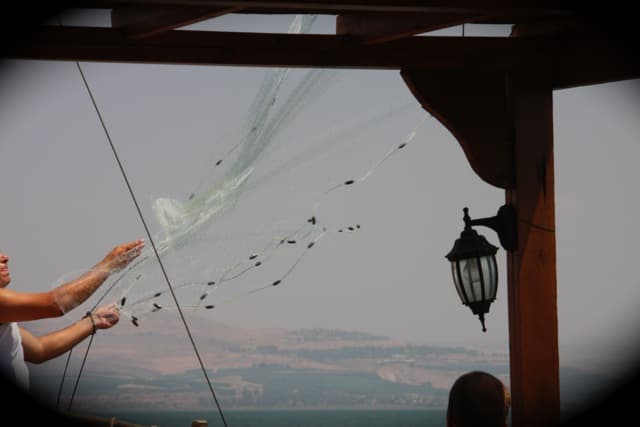 Casting a fishing net from a wooden boat on the Sea of Galilee, hills across the water