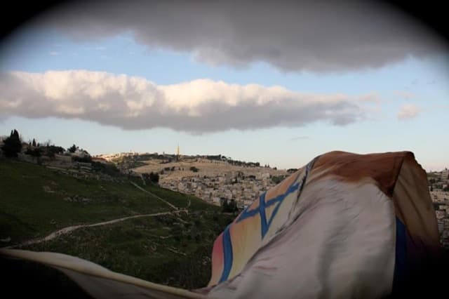 Israeli flag in the foreground above a hillside city at dusk